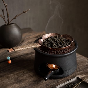 Puede incluir: Un calentador de té de cerámica negra con una abertura para una vela, coronado por una bandeja de metal color cobre en forma de flor llena de hojas de té. El mango está envuelto en cuerda y tiene un pequeño abalorio azul y naranja.