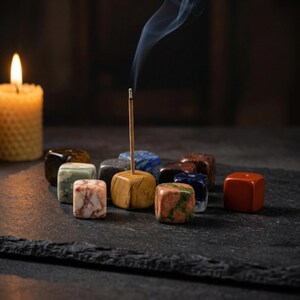 Puede incluir: Una disposici&oacute;n de piedras preciosas coloridas en forma de cubo y una varilla de incienso encendida, con humo ascendente. Una vela encendida est&aacute; en el fondo. Las piedras preciosas son de varios colores.