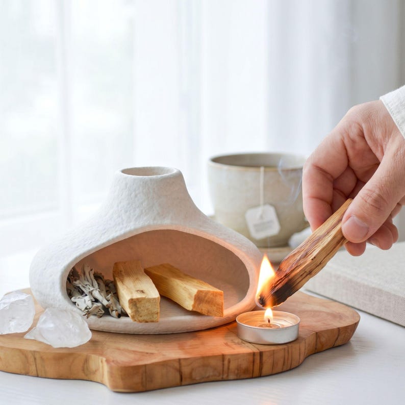 May include: A white ceramic incense holder with a large opening, holding sage and two pieces of palo santo wood. A hand lights a palo santo stick with a small candle on a wooden board. A cup of tea is in the background.