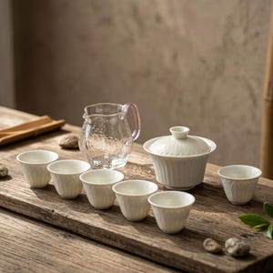 May include: A tea set with a clear glass pitcher with a pink handle, a white lidded tea bowl, and six small white teacups. The set is arranged on a rustic wooden surface, suggesting a traditional tea ceremony.