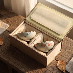 May include: A beige rectangular gift box with a hinged lid containing two small ceramic bowls nestled in fabric lining. One bowl features a blue bamboo leaf pattern, while the other displays a delicate orange floral design on a cream base.