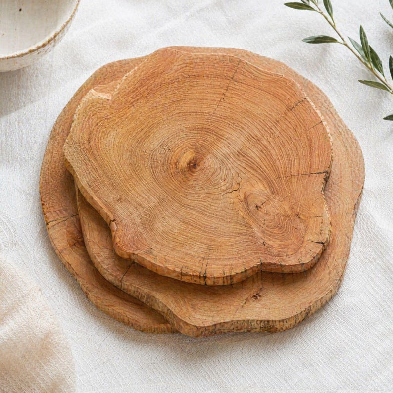 May include: Three wooden, irregularly shaped serving boards with visible wood grain and a light brown color. The boards are stacked on top of each other, with a white cloth in the background. A ceramic bowl is visible in the upper left corner.