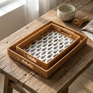 May include: Two rectangular woven trays, one nested inside the other, with a fish pattern on the interior. The trays are light brown and sit on a wooden surface. A ceramic bowl and whisk are in the background.
