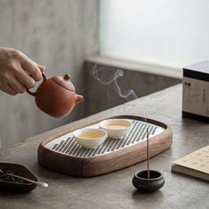 Puede incluir: Una tetera de arcilla de color marrón rojizo con textura, dos tazas de té de cerámica blanca sobre una bandeja de madera rectangular con un inserto de metal perforado, un pequeño porta-incienso oscuro y un plato de madera con hojas de té.