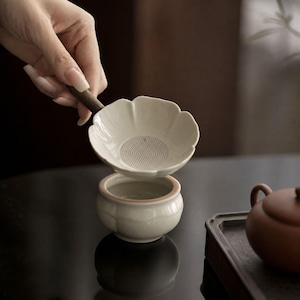 May include: A cream-colored ceramic tea strainer shaped like a flower with a dark wooden handle, positioned above a matching cream-colored ceramic tea cup with a rounded base and a subtle scalloped rim design.