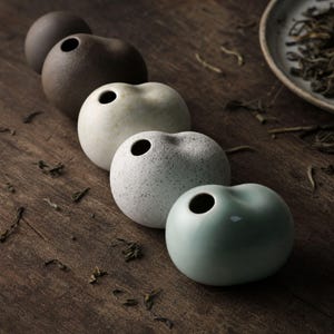 May include: A collection of five small, rounded ceramic vases arranged in a diagonal line. The vases feature varied textures and finishes, including matte dark brown, speckled off-white, textured grey, and a smooth, glossy pale green glaze.