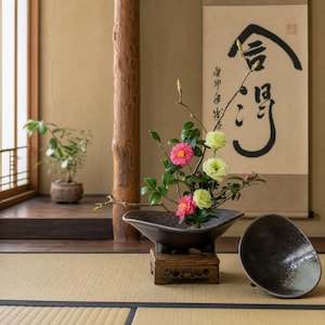 May include: An Ikebana floral arrangement featuring pink and yellow flowers in a dark ceramic bowl, set on a wooden stand. A second, matching bowl sits nearby. The background includes a scroll with Japanese calligraphy and a small potted plant.