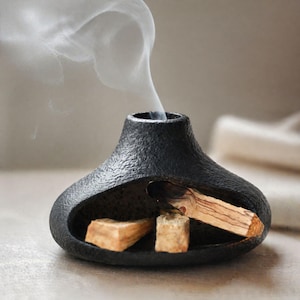 May include: A black, textured ceramic incense holder with a unique shape. The holder contains burning palo santo wood, emitting wisps of white smoke. Two additional pieces of palo santo are visible inside the holder.