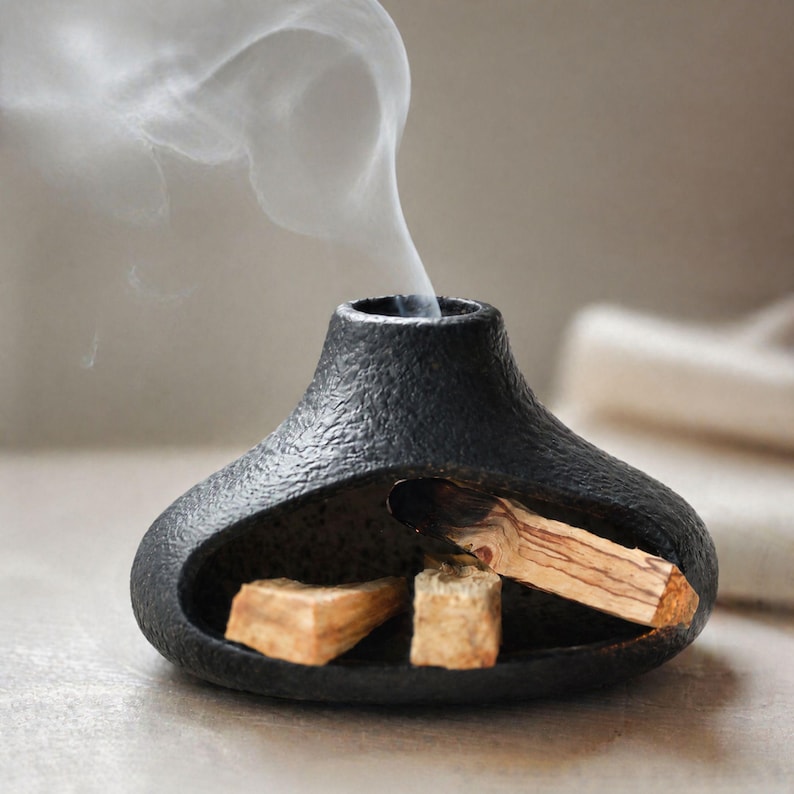 May include: A black, textured ceramic incense holder with a unique shape. The holder contains burning palo santo wood, emitting wisps of white smoke. Two additional pieces of palo santo are visible inside the holder.