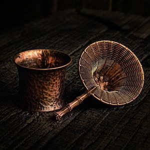 Puede incluir: Una taza de cobre con textura martillada y un colador de cobre tejido con un mango estilo bambú. Los objetos se exhiben sobre una superficie de madera oscura, mostrando una estética rústica.