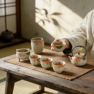 May include: A tea set with a floral design. The set includes a teapot, a creamer, a small incense burner, and several cups. The cups have a unique petal shape. The set is placed on a brown mat on a wooden table.
