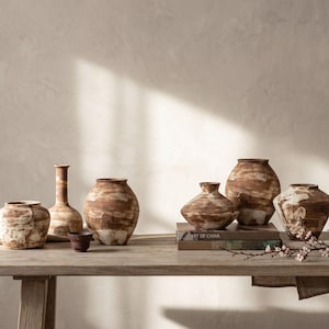 May include: A collection of six ceramic vases and one small bowl featuring a rustic, marbled brown and white finish. The vessels vary in shape, including rounded, tapered, and wide-mouthed designs, arranged on a wooden surface with a book titled ART OF CHINA.