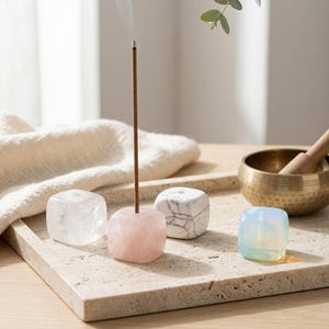 Puede incluir: Conjunto de portainciensos c&uacute;bicos en varios colores: transparente, rosa, blanco y azul. Un palito de incienso encendido en el soporte rosa, con humo ascendente. Un cuenco tibetano de lat&oacute;n y una maza de madera en el fondo.