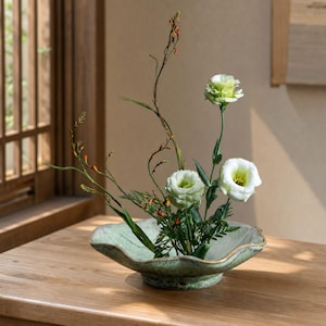 Peut inclure: Un bol en céramique peu profond à bords festonnés avec un vernis vert clair moucheté contenant un arrangement floral. L'arrangement comprend deux fleurs d'eustoma blanches, du feuillage vert et de longues tiges fines à bourgeons orangés.