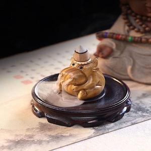 Puede incluir: Un quemador de incienso de madera con forma de hipopótamo, con un sombrero pequeño. El hipopótamo está sobre un soporte de madera oscura, con humo saliendo de su boca. El fondo presenta una figura borrosa y una superficie estampada.