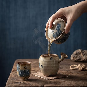 Könnte beinhalten: Eine Keramik-Teekanne mit seitlichem Griff und passendem Deckel gießt dampfenden Tee in eine kleine Keramiktasse. Beide Teile haben eine gesprenkelte beige Glasur mit dunkelblauen rechteckigen Feldern und kleinen weißen Blumenmustern.
