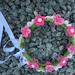 Puede incluir: Una corona floral hecha a mano con flores rosas y blancas, hojas verdes y una cinta blanca. La corona está hecha de un material suave y difuso, ideal para una boda o festival. La corona se encuentra sobre un fondo de rocas grises.