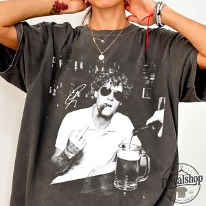 May include: A charcoal gray short-sleeve t-shirt featuring a black and white graphic print of a person wearing sunglasses, smoking a cigar, and gesturing with a middle finger while a hand pours a drink into a glass mug. The shirt has a crew neckline.