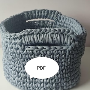 May include: A gray, hand-crocheted basket with two handles. The basket is made of thick, soft yarn and has a textured appearance. The word "PDF" is in a white speech bubble on the side of the basket. The basket is likely for storage or decoration.