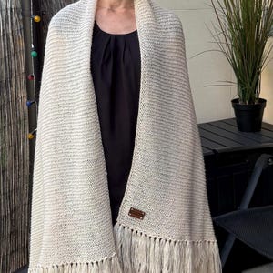 May include: A cream-colored knitted shawl featuring a textured stitch pattern and long fringe along the bottom edge. A small rectangular brown leather-like tag is attached to the lower front section of the knit fabric, providing a subtle contrast.
