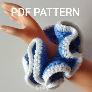 Puede incluir: Una scrunchie de ganchillo azul y blanco con el texto "PDF PATTERN" en blanco. La scrunchie está hecha de un hilo suave y tiene un diseño con volantes. La scrunchie tiene aproximadamente 10 cm de diámetro.