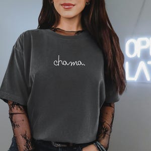 Puede incluir: Camiseta gris oscuro con la palabra "chama" en escritura blanca. La camiseta tiene mangas cortas y un aspecto ligeramente descolorido. La modelo lleva una camisa negra de manga larga de encaje debajo.
