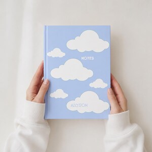 Puede incluir: Un cuaderno de tapa dura de color azul claro con un patrón de nubes blancas. La palabra NOTES está impresa en blanco cerca del centro superior, y el nombre ALLYSON está impreso en azul claro sobre una nube blanca grande en la parte inferior.