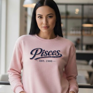 Puede incluir: Sudadera rosa claro con la palabra "Pisces" en escritura azul marino con contorno blanco y detalles rojos. Debajo, el texto dice "EST. 1988". La sudadera es de cuello redondo.
