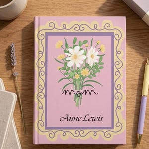 Puede incluir: Un diario rosa con un diseño floral y la palabra "mamá" en cursiva. La portada presenta un ramo de flores blancas y amarillas, enmarcado por un borde decorativo. El nombre "Anne Lewis" está impreso debajo.