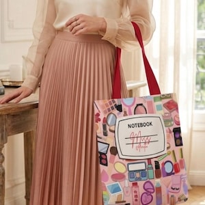 Puede incluir: Un bolso tote rosa con asas rojas, decorado con ilustraciones de maquillaje. El bolso presenta la palabra "NOTEBOOK" y un espacio para un nombre. El bolso lo sostiene una persona que lleva una falda plisada rosa y una blusa transparente.