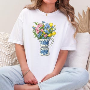Puede incluir: Camiseta blanca con diseño floral. El gráfico muestra una cafetera llena de un ramo de flores coloridas en tonos rosa, azul y amarillo. La cafetera tiene un estampado floral azul.