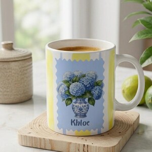 Op de afbeelding: Een witte keramische mok met een verticaal geel en wit gestreept patroon. Het midden toont een lichtblauwe rechthoekige afbeelding met een geschulpte rand, met een blauw-witte vaas gevuld met blauwe hortensia's en de naam Khloe.