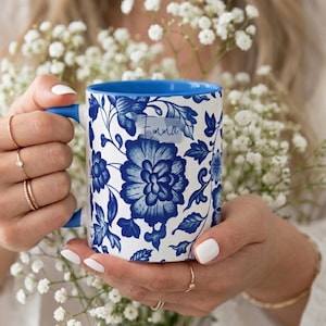 Puede incluir: Taza de cerámica blanca con interior y asa azules, con un diseño floral azul. La taza tiene el nombre "Emma" impreso. La taza se sostiene frente a un fondo de flores blancas.