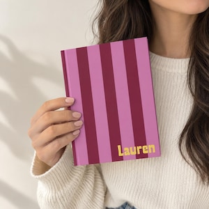 Puede incluir: Un cuaderno de tapa dura con un patrón de rayas verticales en tonos alternos de burdeos oscuro y rosa claro. El nombre Lauren está impreso en una fuente amarilla negrita sin remates en la esquina inferior derecha de la portada.