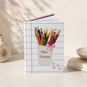 Puede incluir: Un cuaderno blanco con temática escolar. La portada presenta una ilustración de un vaso de lápices con lápices de colores, un lazo rosa y el texto "Miss. Emma". El cuaderno tiene páginas rayadas y un lomo con estampado rosa.
