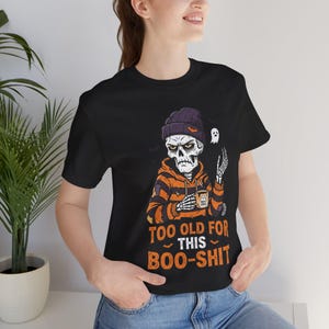 May include: A black short-sleeve t-shirt featuring a graphic of a skeleton wearing a purple beanie and an orange and black striped hoodie, holding a mug. The text reads, "TOO OLD FOR THIS BOO-SHIT" in orange letters below the skeleton illustration.