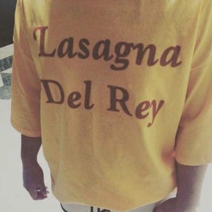 Peut inclure: T-shirt jaune à manches courtes avec le texte "Lasagna Del Rey" en script marron. Le t-shirt est porté avec un short blanc à bordure noire. La personne tient un téléphone.