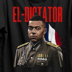 May include: Black t-shirt featuring a portrait of a man in a military uniform with the text "EL-DICTATOR" in red. A French flag is visible in the background. The shirt is hanging on a black hanger.