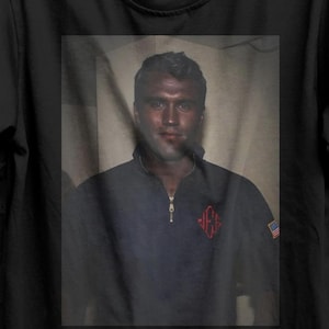 May include: Black t-shirt featuring a framed image of a person in a navy blue zip-up jacket with a red emblem. The shirt also displays the text "SEVENTHREESEVEN" and a "BUY 3 FREE" graphic.