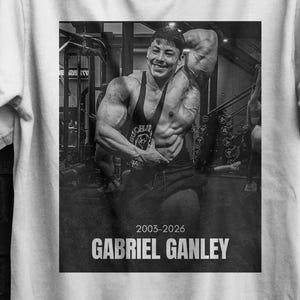 Könnte beinhalten: Weißes T-Shirt mit einem Schwarz-Weiß-Bild eines Bodybuilders, dem Text "GABRIEL GANLEY" und den Jahren "2003-2026". Das Shirt hat auch den Text "SEVENTHREESEVEN" und eine gelb-rosa Flugzeuggrafik.