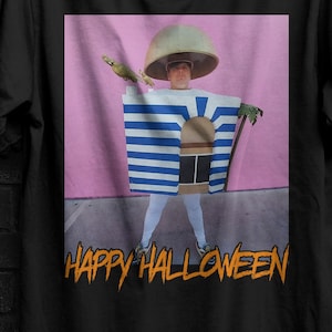 Puede incluir: Una camiseta negra de manga corta con un estampado gráfico de una persona con un sombrero en forma de seta y un disfraz de caja a rayas azules y blancas. El gráfico incluye el texto "HAPPY HALLOWEEN" en letras naranjas en la parte inferior.