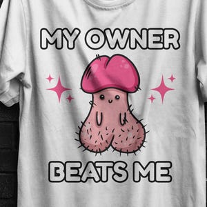May include: A white short-sleeved t-shirt featuring a cartoon illustration of a pink phallic character with a smiling face and small black eyes. The text MY OWNER BEATS ME is printed in black capital letters above and below the illustration.