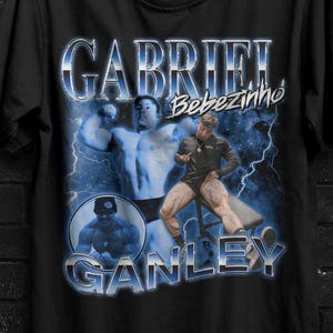 Könnte beinhalten: Schwarzes T-Shirt mit einem Retro-Grafik eines Mannes, dem Namen "Gabriel" und "Ganley" in Blau. Das Shirt enthält auch den Text "Bebezinho". Eine gelb-schwarze "Buy 3 Free" Grafik befindet sich oben rechts.