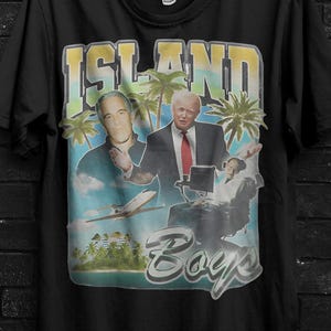 May include: Black t-shirt featuring a graphic design with the text "ISLAND BOYS" and images of two men, palm trees, and a tropical scene. The shirt also has the text "BUY 3 FREE" and "CODE: 3SHIP".