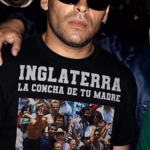 Op de afbeelding: Een zwart t-shirt met de witte tekst "INGLATERRA LA CONCHA DE TU MADRE" boven een grafische print van een menigte, met een persoon in een blauw-wit voetbalshirt en een ander persoon die een handgebaar maakt.
