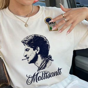 Può includere: Una t-shirt a maniche corte color crema con un'illustrazione grafica nera di un uomo che fuma una sigaretta di profilo. Sotto la grafica è presente il testo Moltisanti in carattere corsivo. Una piccola toppa rettangolare con il testo Christopher Moltisanti e una bandiera italiana è posizionata sulla parte superiore del petto.