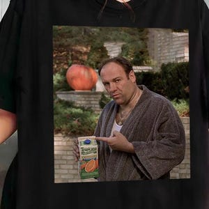 Può includere: Una t-shirt nera con una stampa grafica di un uomo in accappatoio marrone che indica un cartone di succo d'arancia Tropicana. L'immagine mostra un muro di mattoni e vegetazione sullo sfondo. La maglietta ha un girocollo e maniche corte.