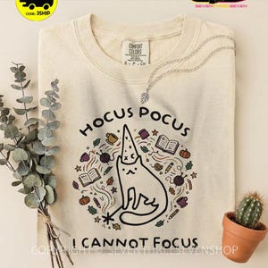 Könnte beinhalten: Ein elfenbeinfarbenes T-Shirt mit einer Grafik einer Katze, die einen Zaubererhut trägt und einen Zauberstab hält. Der Text lautet HOCUS POCUS I CANNOT FOCUS. Um die Katze herum sind Illustrationen von Büchern, Kürbissen, Garnknäueln und Blättern.