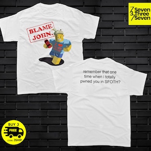 Op de afbeelding: Wit t-shirt met een afbeelding van een stripfiguur met de tekst "BLAME JOHN." De achterkant van het shirt heeft de tekst "remember that one time when i totally pwned you in SFOTH?"