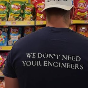 Könnte beinhalten: Eine weiße Baseballkappe mit dem Text MAKE EUROPE GREAT AGAIN in schwarzen Buchstaben und ein dunkelblaues T-Shirt mit dem weißen Text WE DON'T NEED YOUR ENGINEERS auf dem Rücken, vor Regalen mit verschiedenen Tüten Lay's Kartoffelchips.